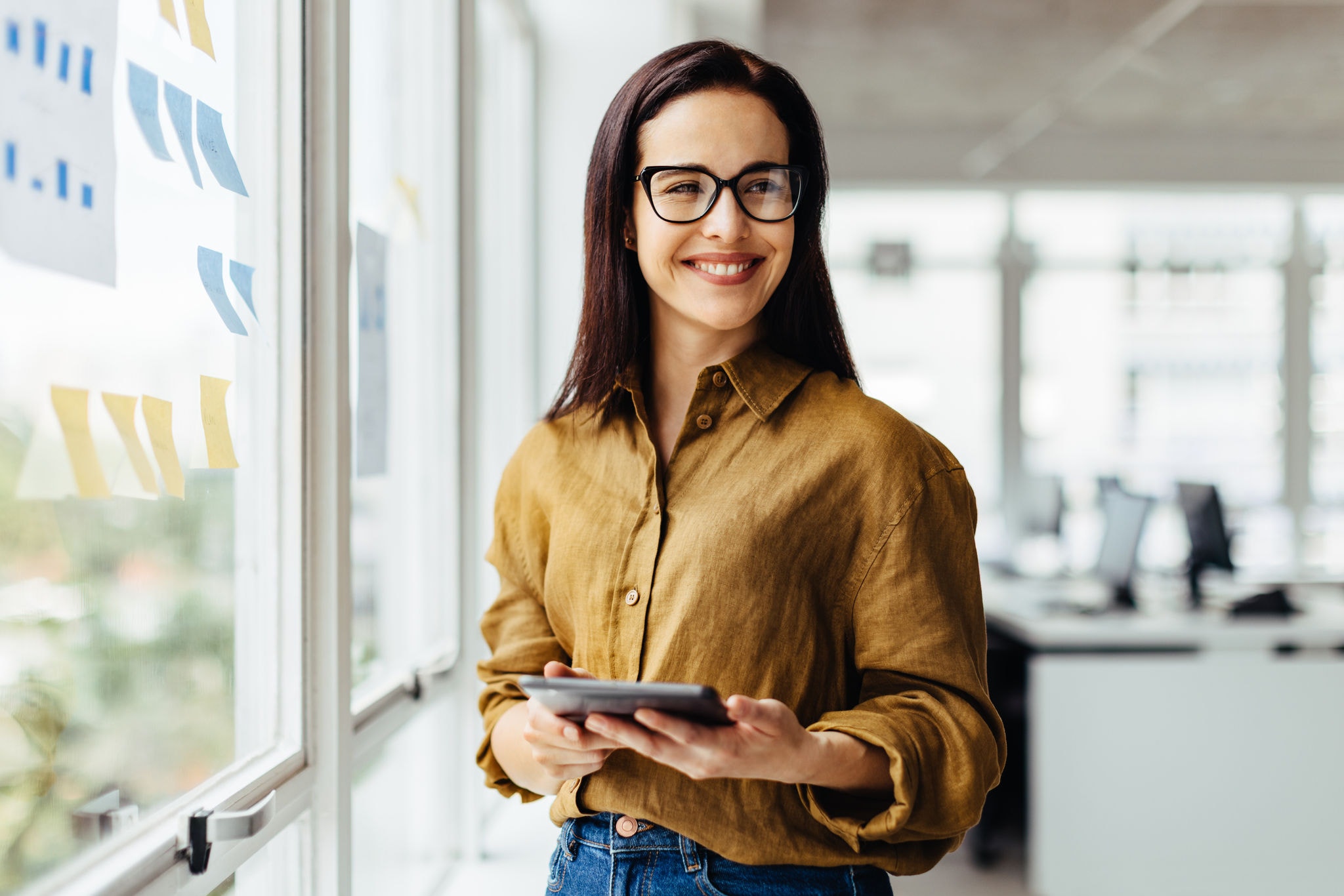 Thoughtful business woman holding a tablet while standing next to a window. Happy young business woman working for an advertising agency.