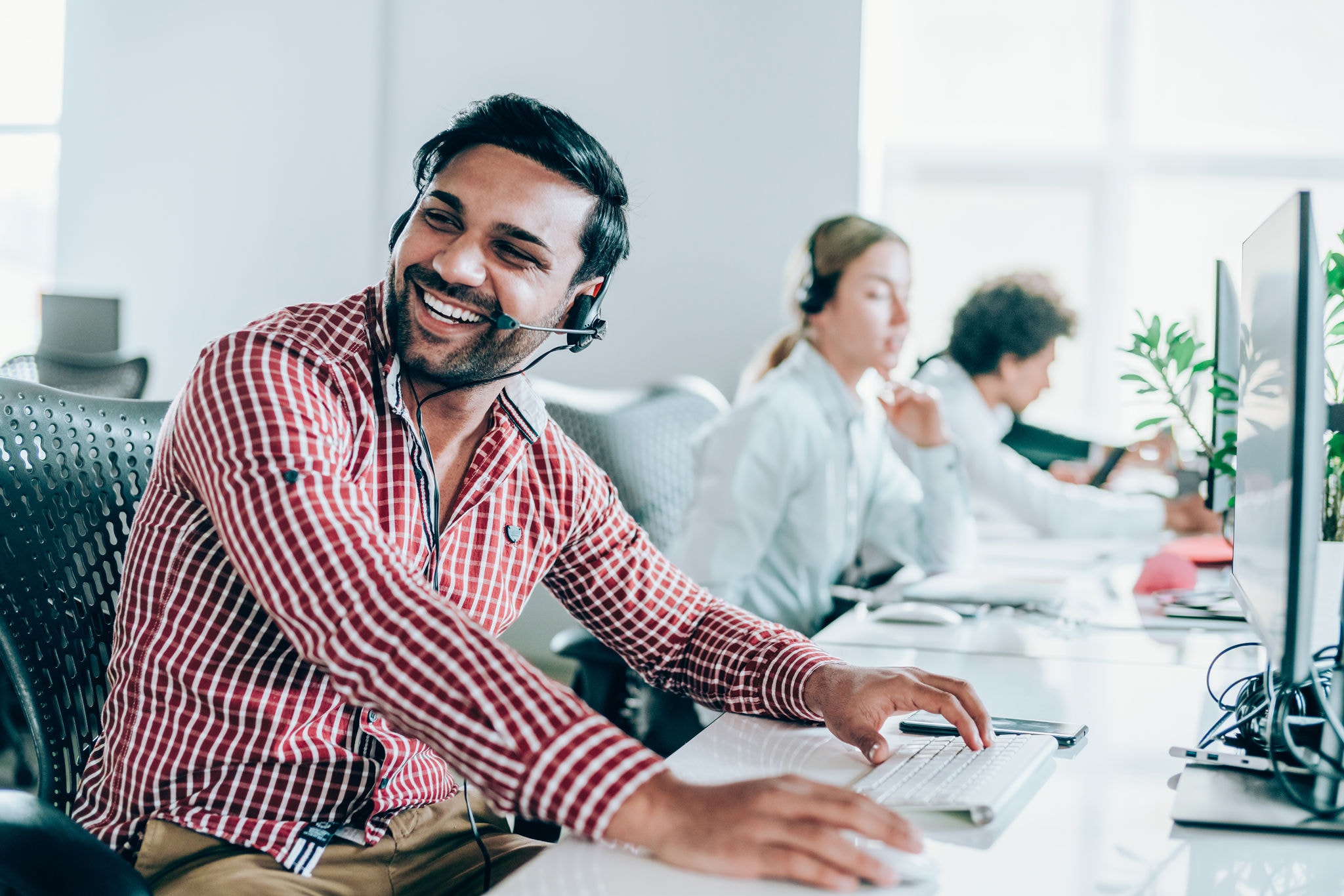 Smiling handsome ethnicity businessman working in call center. Shot of a cheerful young man working in a call center with his team. Confident male operator is working with colleagues. Call center operators sitting in a row at desks.