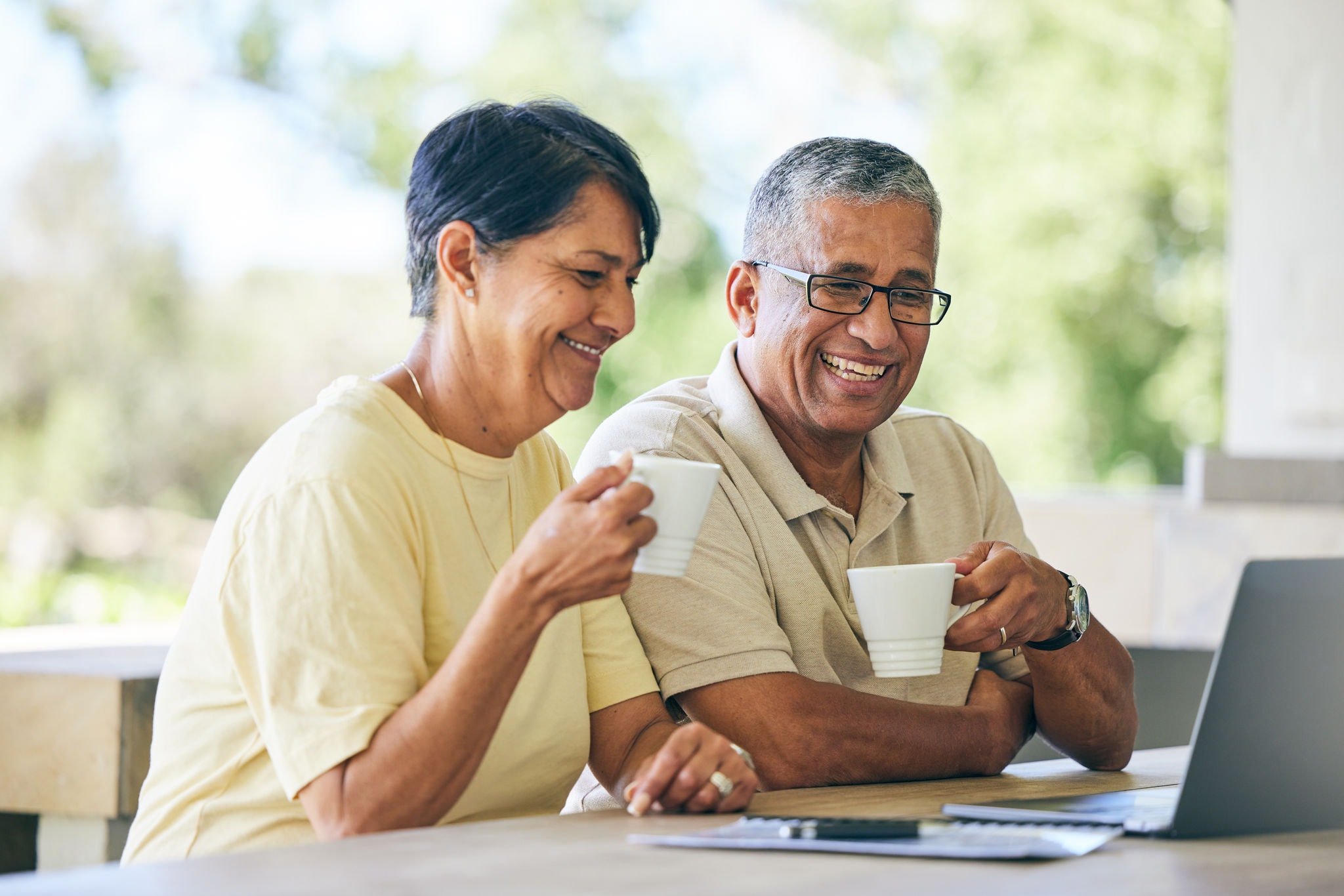 Elderly couple enjoying coffee and laughing together while watching something on their laptop.