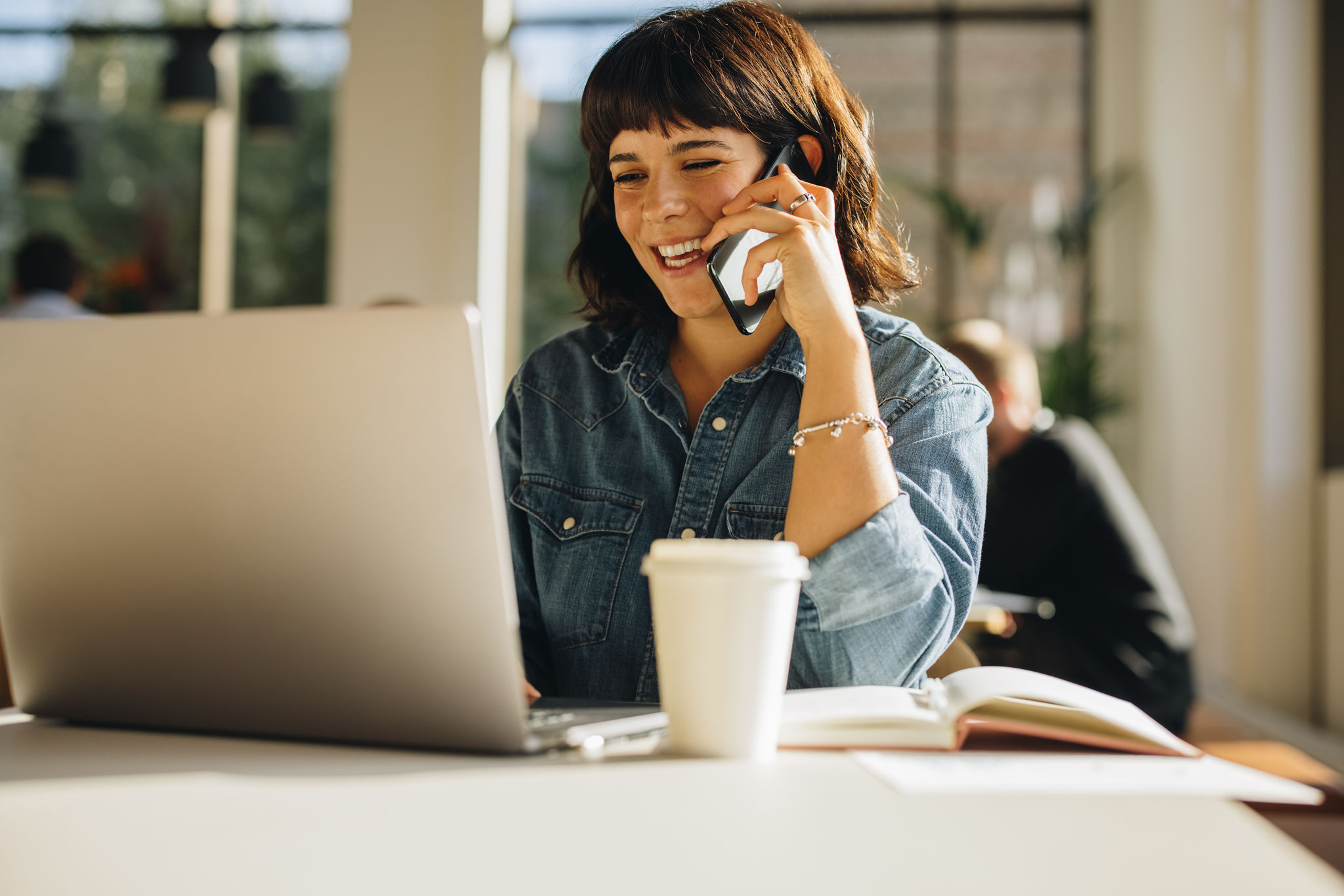 Cheerful businesswoman talking on cell phone while using laptop. Smiling young woman speaking on smart phone while looking at laptop in a coworking space.