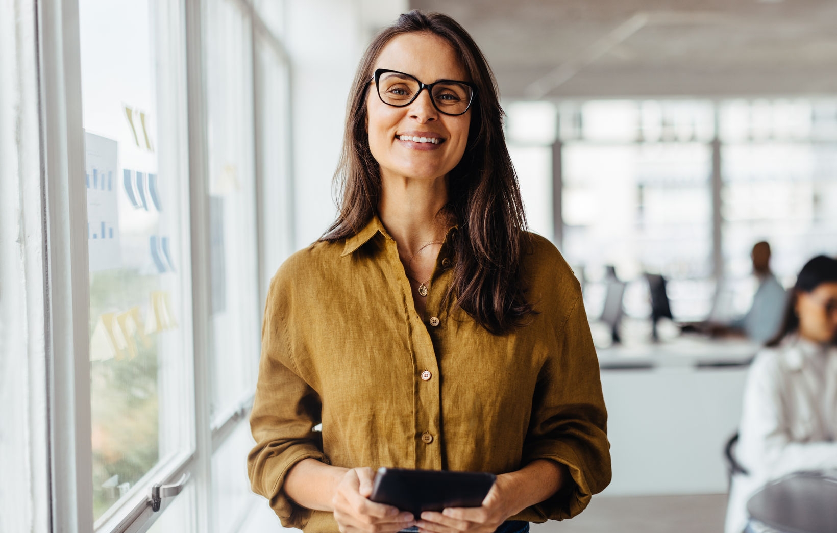 Business woman smiling at the camera while holding a tablet pc. Mature business woman standing in an office with her colleagues working in the background.