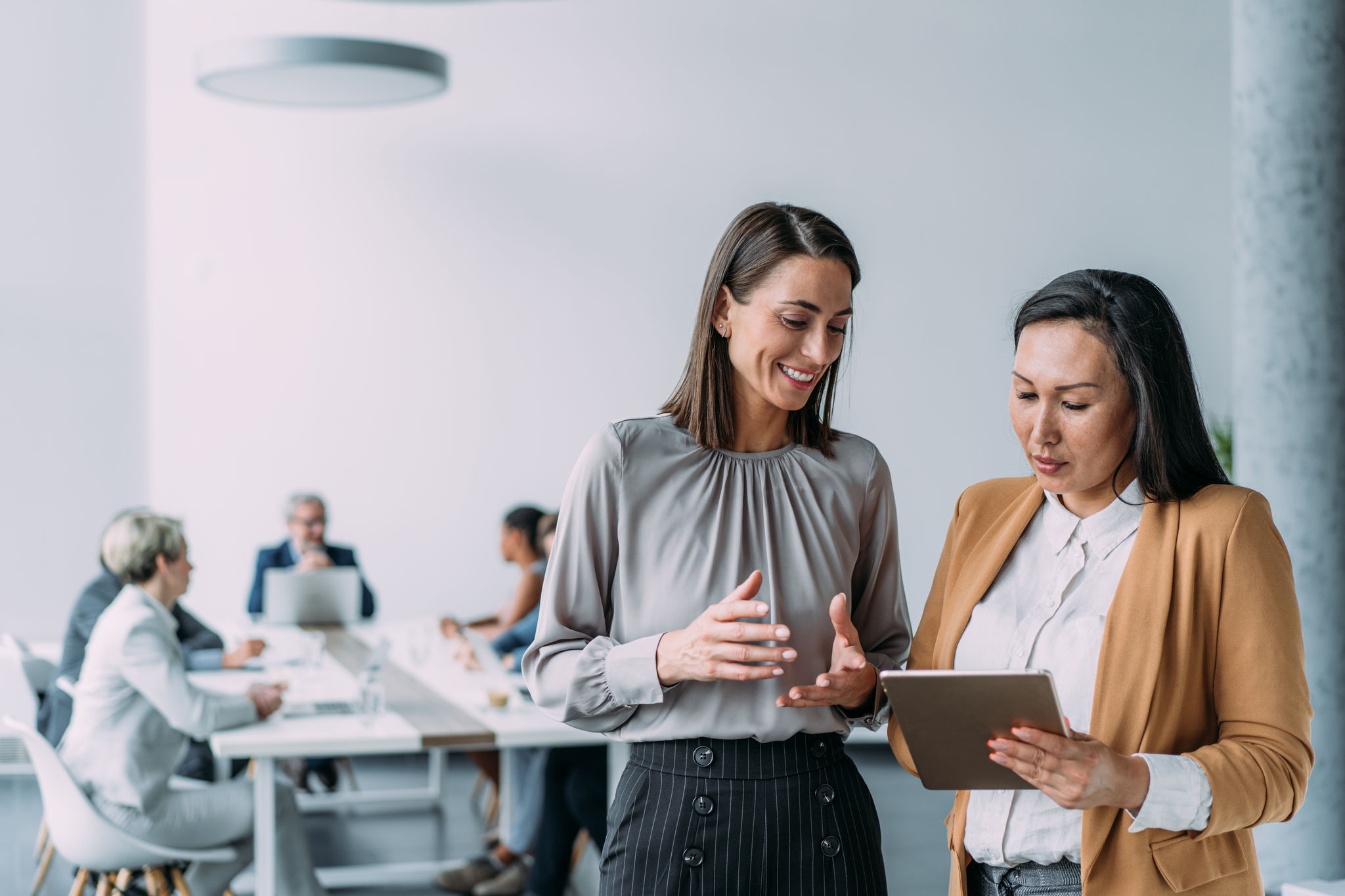 Shot of two female coworkers having a discussion in modern office. Businesswomen in meeting using digital tablet and discussing business strategy. Confident business people working together in the office. Corporate business persons discussing new project and sharing ideas in the workplace. Successful businesswomen standing with their colleagues working in background.