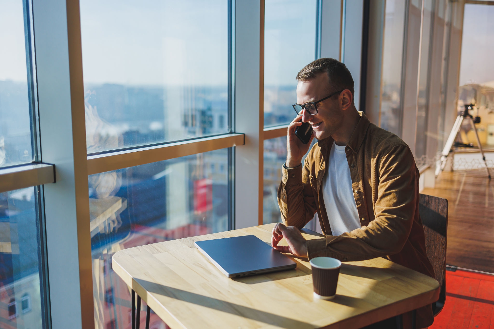 A happy young man wearing glasses and casual clothes is talking on the phone while working on a laptop from a cozy workspace. A successful freelancer works remotely