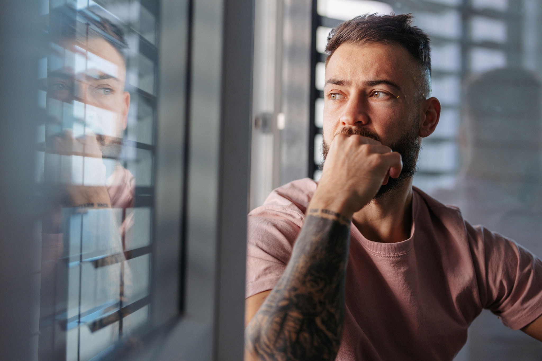 Handsome calm man in pink t-shirt with tattoo on arms looking through window.
