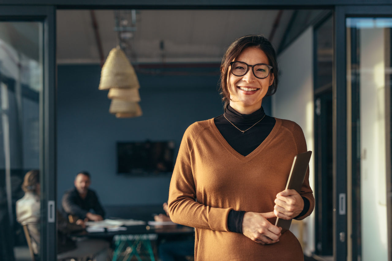Portrait of positive female executive holding a laptop standing in office with colleagues working in background. Smiling business woman in casuals at office.