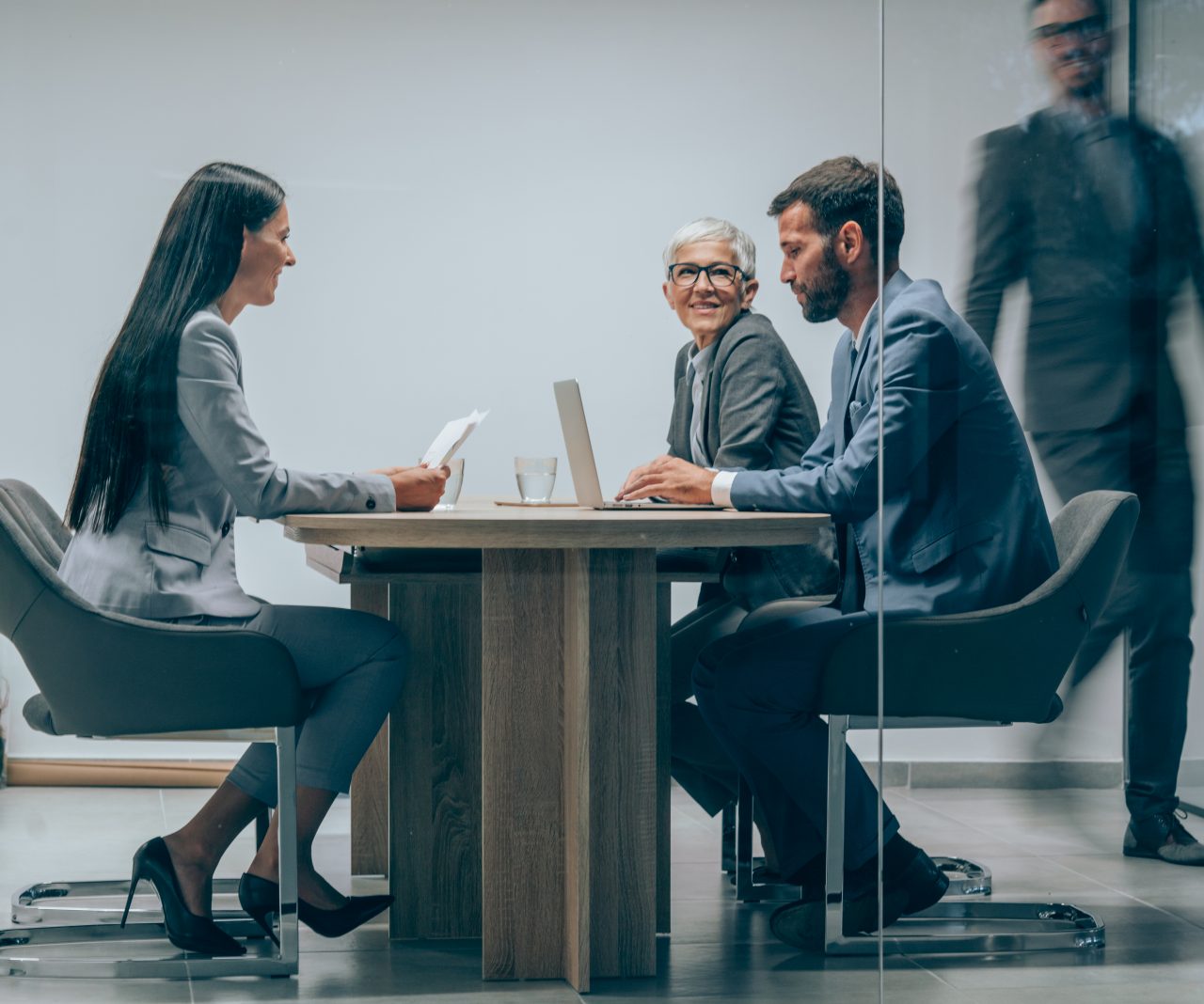 Shot of a  group of business people having a meeting in board room. Businessman walking in blurred motion around his colleagues. Group of entrepreneurs on meeting in board room. Corporate business team on meeting in the office. The view is through glass.
