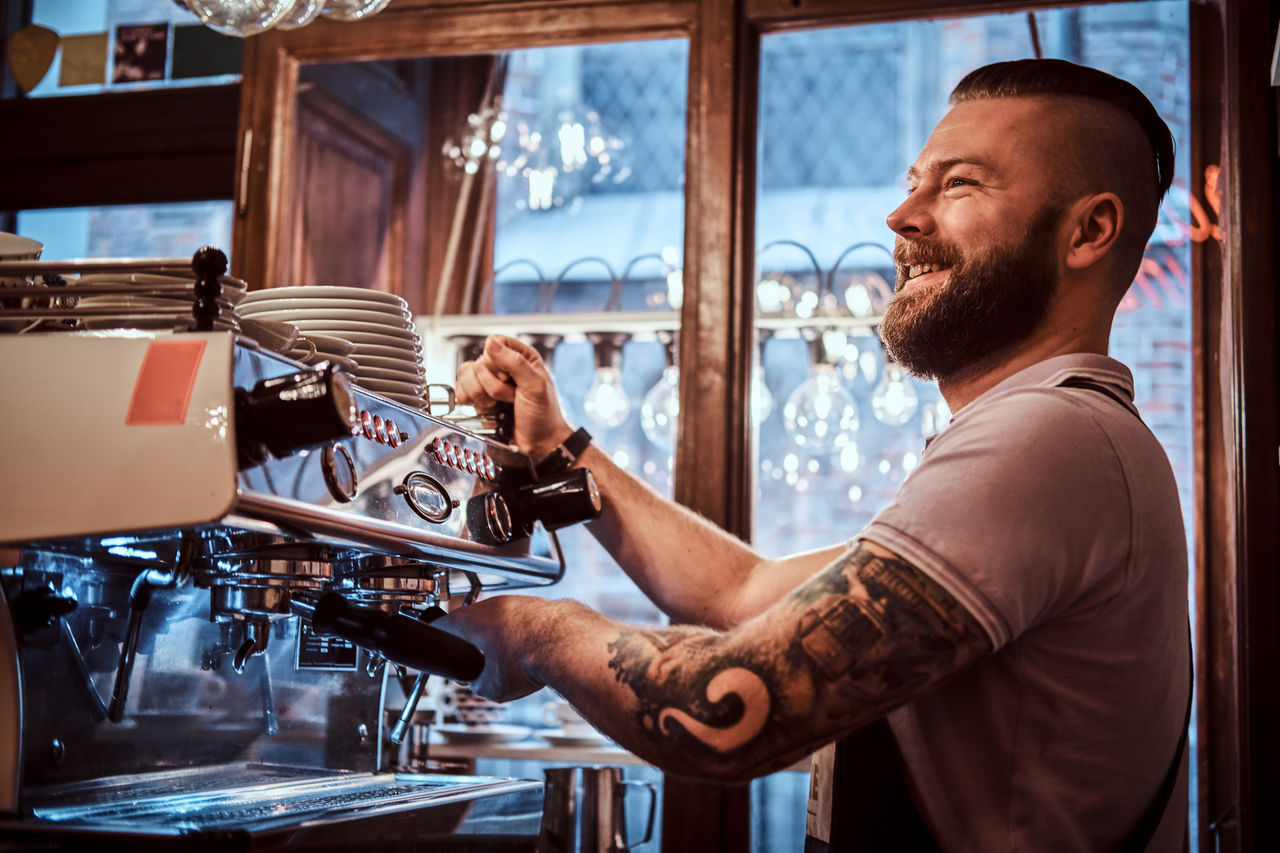 Cheerful barista with stylish beard and hairstyle making coffee for a customer in the coffee shop