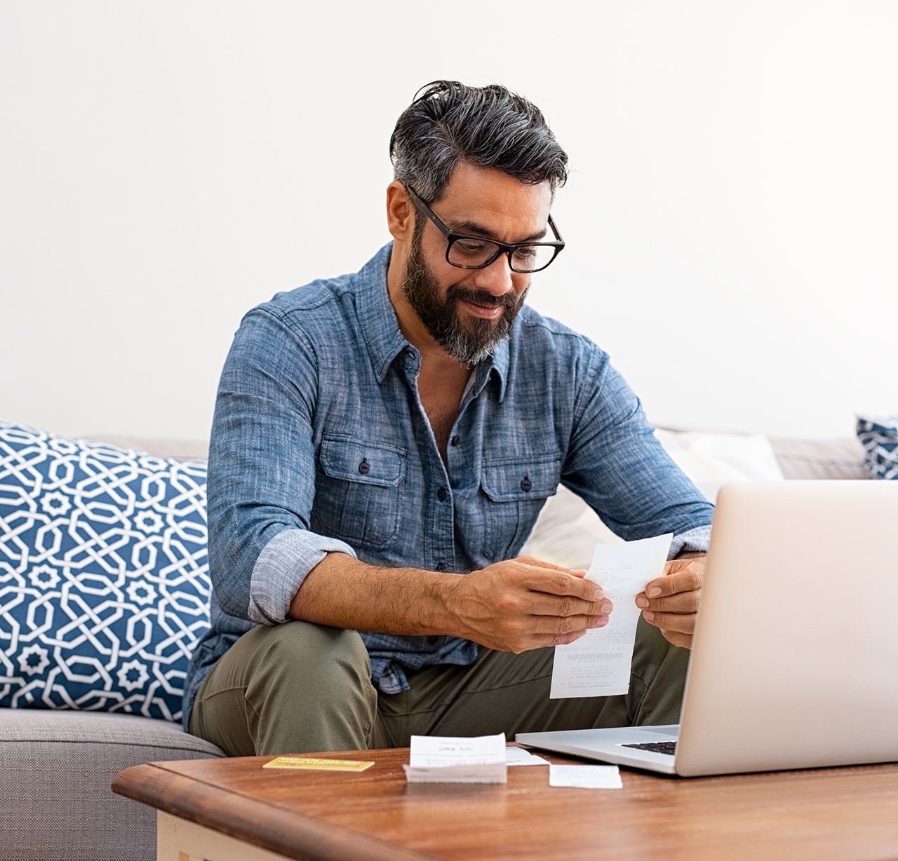 Mature casual man using laptop while looking at invoice. Smiling latin man managing finance with bills and laptop while sitting on couch at home. Hispanic guy reading expenses wearing eyeglasses.