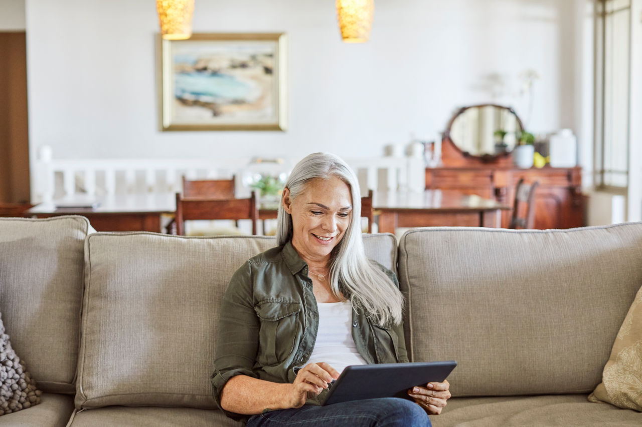 Mature woman smiling while using digital tablet. Happy female is sitting on sofa in living room. She is at home.