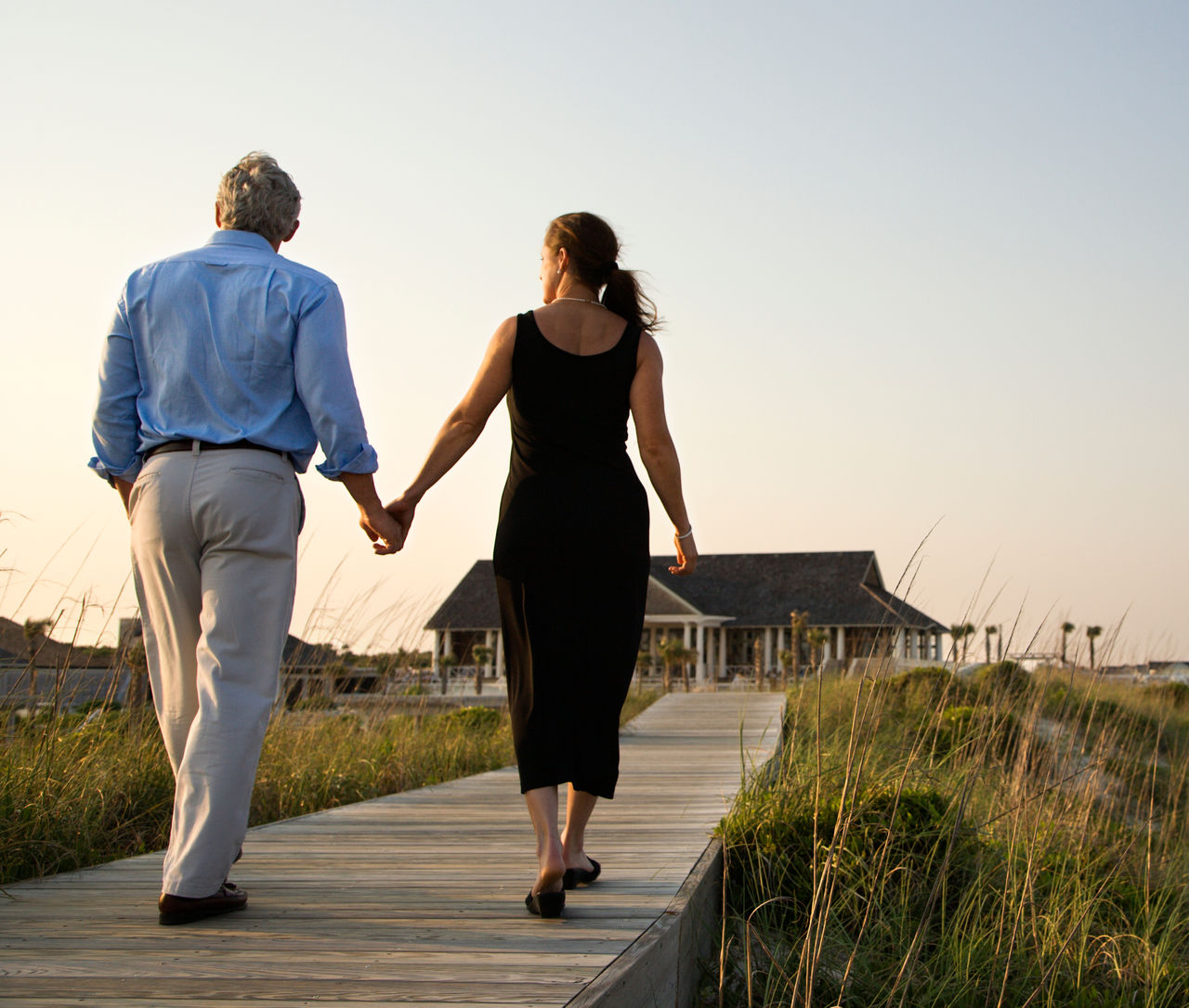 Couple walk hand in hand on a boardwalk towards a beach pavilion. Horizontal shot.