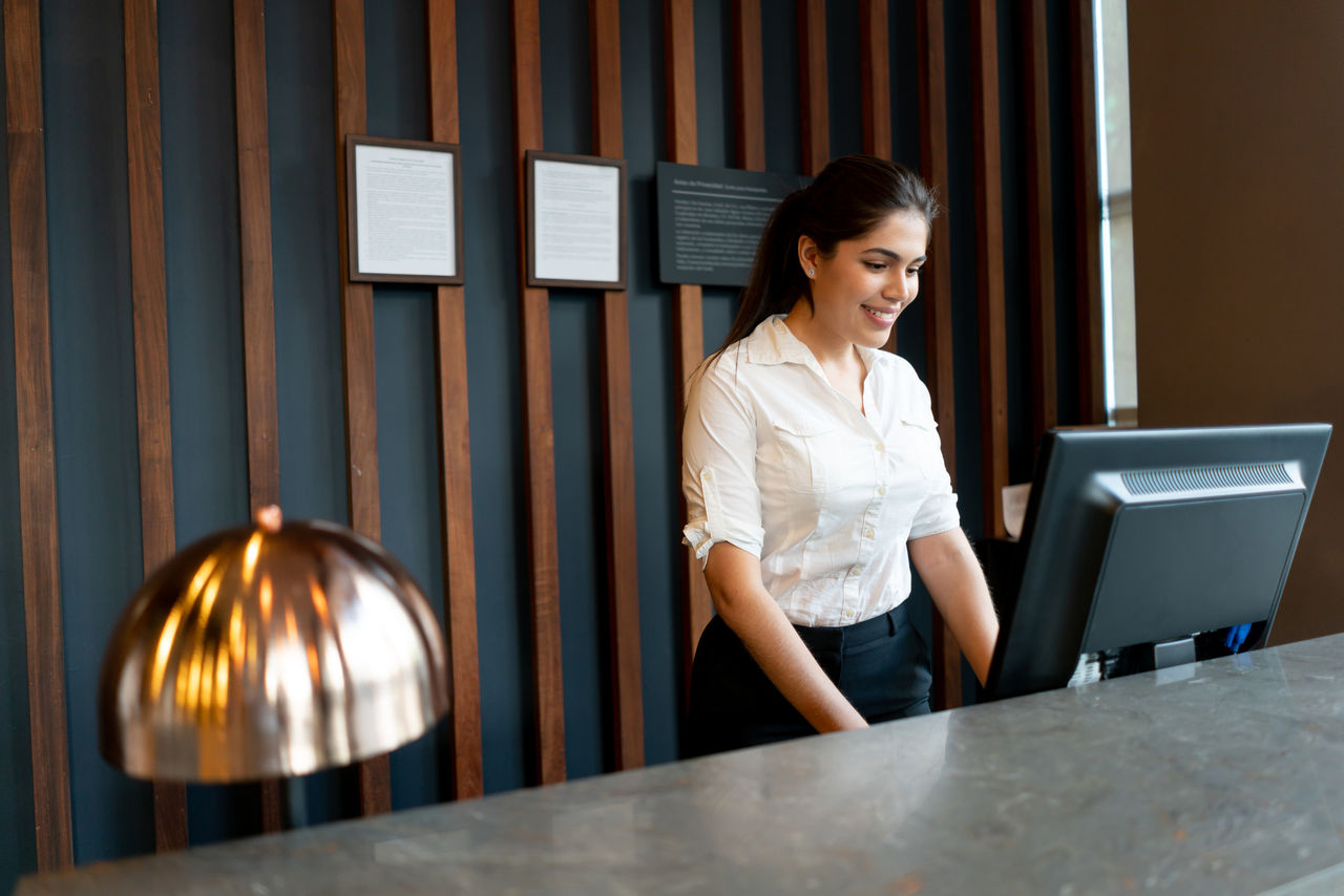 Latin american hotel receptionist working behind counter smiling very happy while looking at computer screen