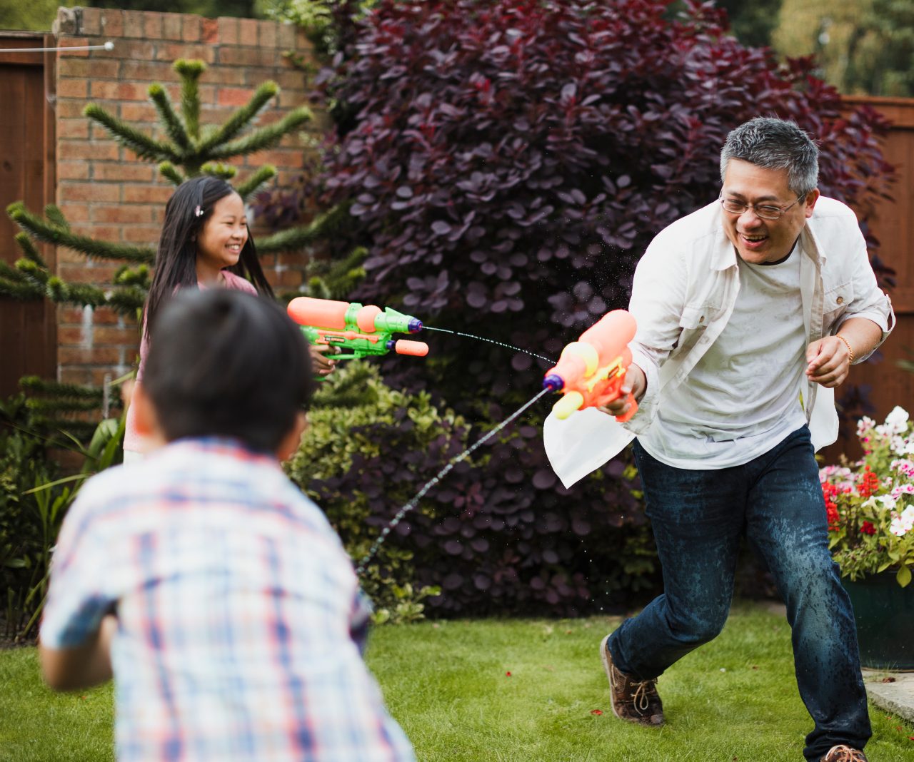 Family are having a water fight together with water pistols in the garden. The little girl is aiming for her dad, who is aiming for the little boy.