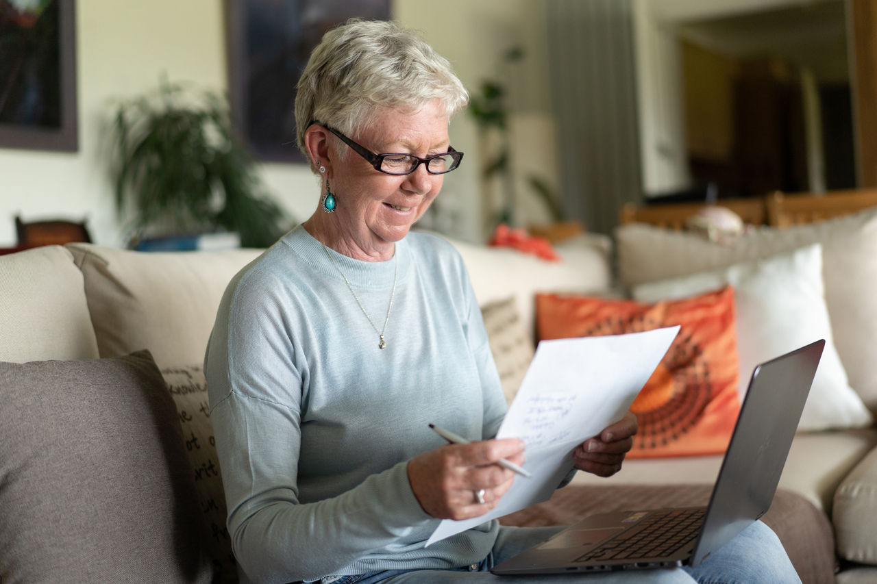 A smiling grey haired lady is studying the numbers for her financial future, while her laptop is on her knees, sitting at home on a sofa in a living room