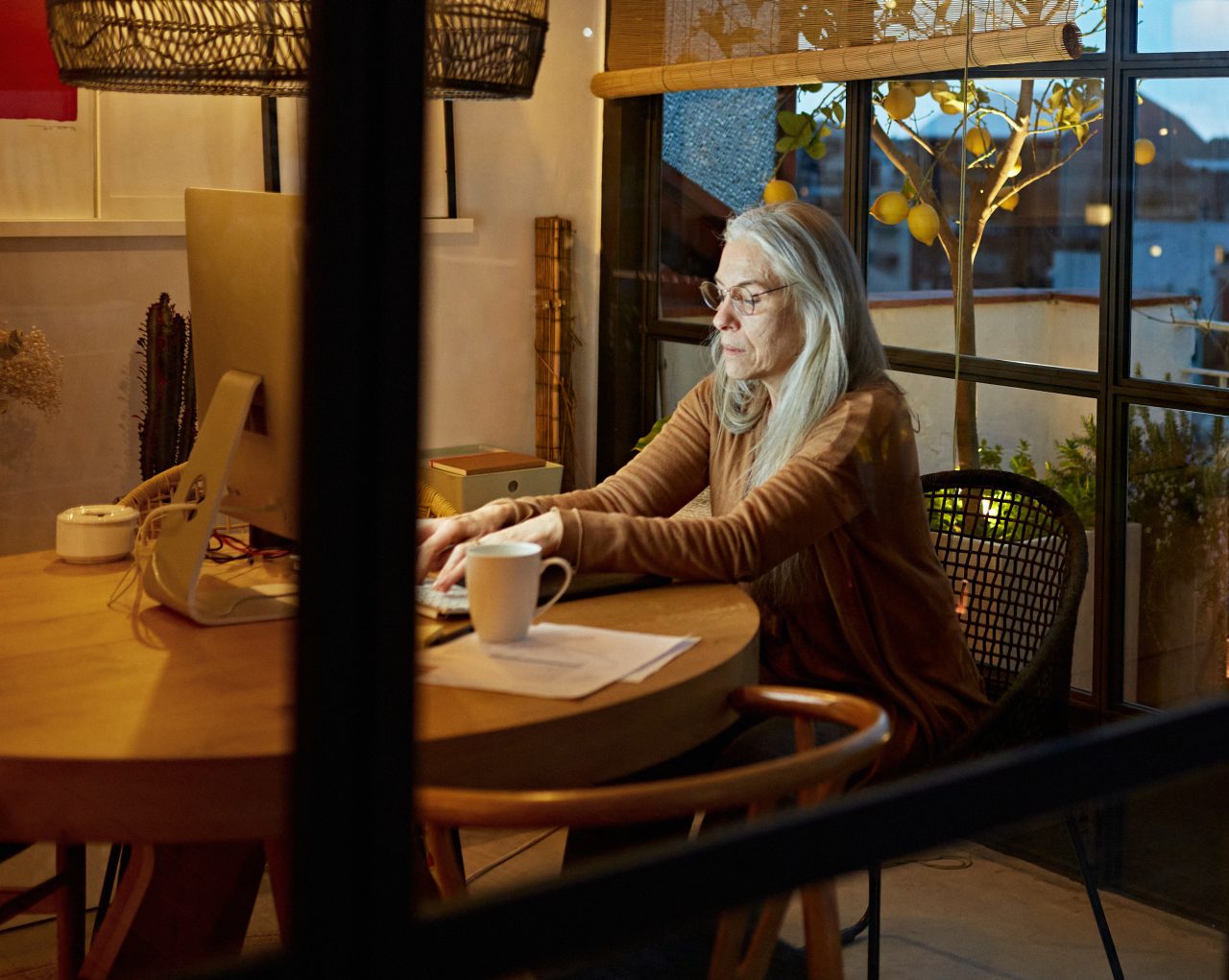 Partial view of gray-haired Caucasian woman wearing eyeglasses and casual clothing photographed through sun room window at end of workday.