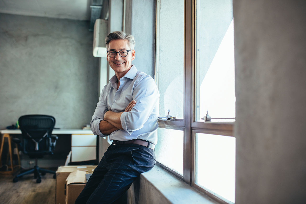 Smiling businessman looking at camera as he stands by a window sill with arms crossed. Mid adult businessman at his office.