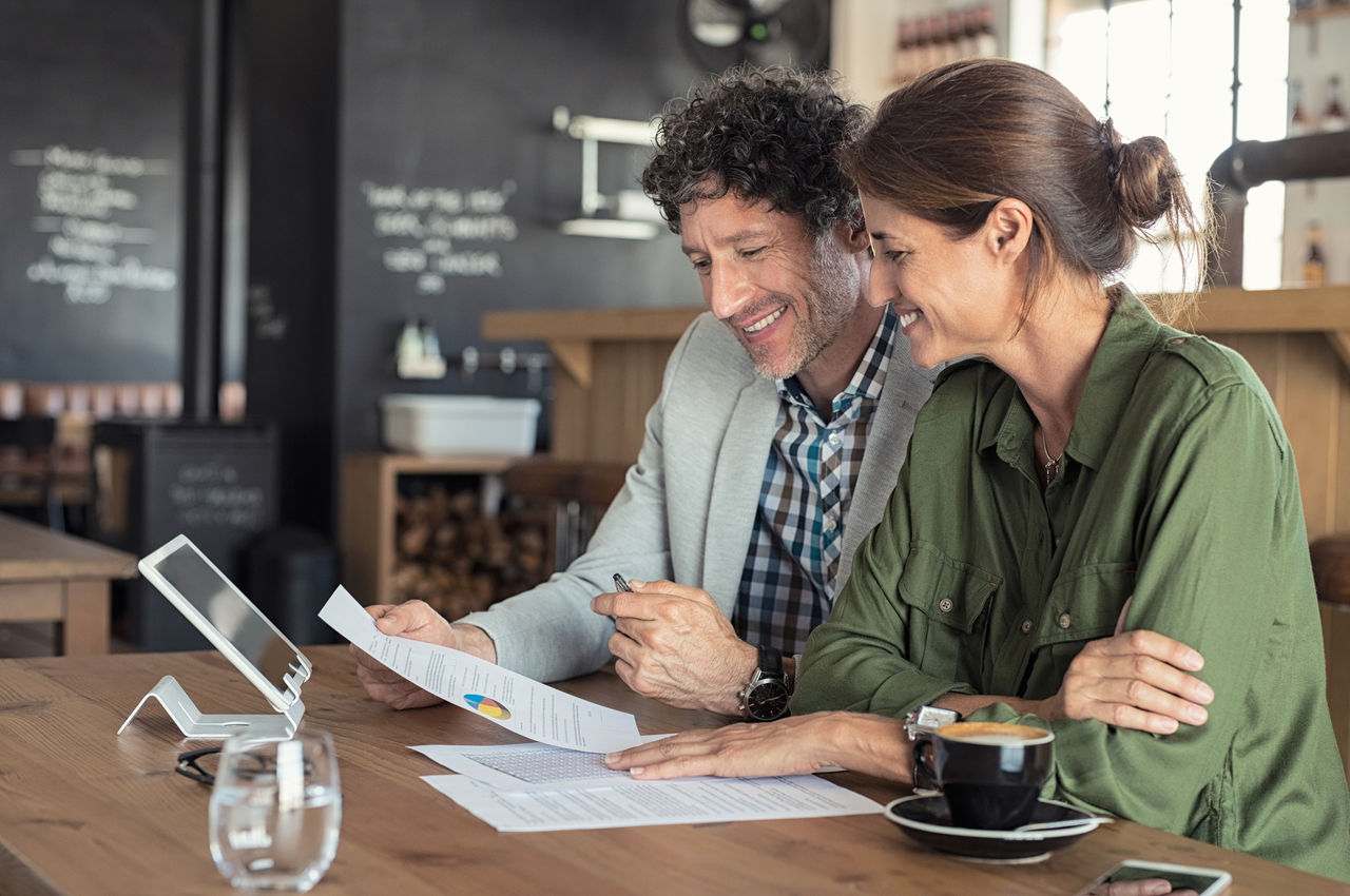 Portrait of mature business man and casual businesswoman sitting in cafe and discussing sales graph. Group of two middle aged coworkers working comparing forecasting graphics. Happy businessman reading report with his business partner.