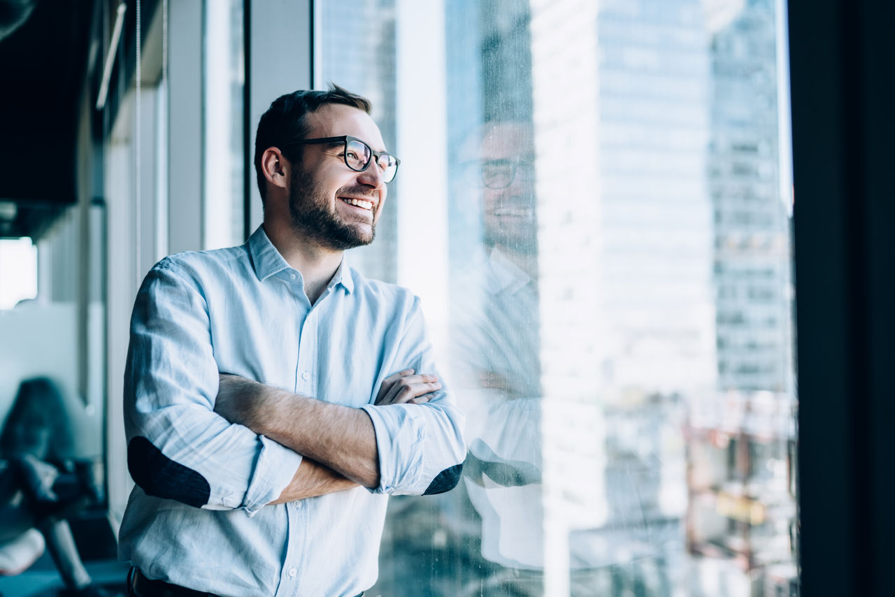 Cheerful male entrepreneur with crossed hands standing near office window view and smiling during work day in company, Caucasian successful corporate boss feeling good from wealthy lifestyle