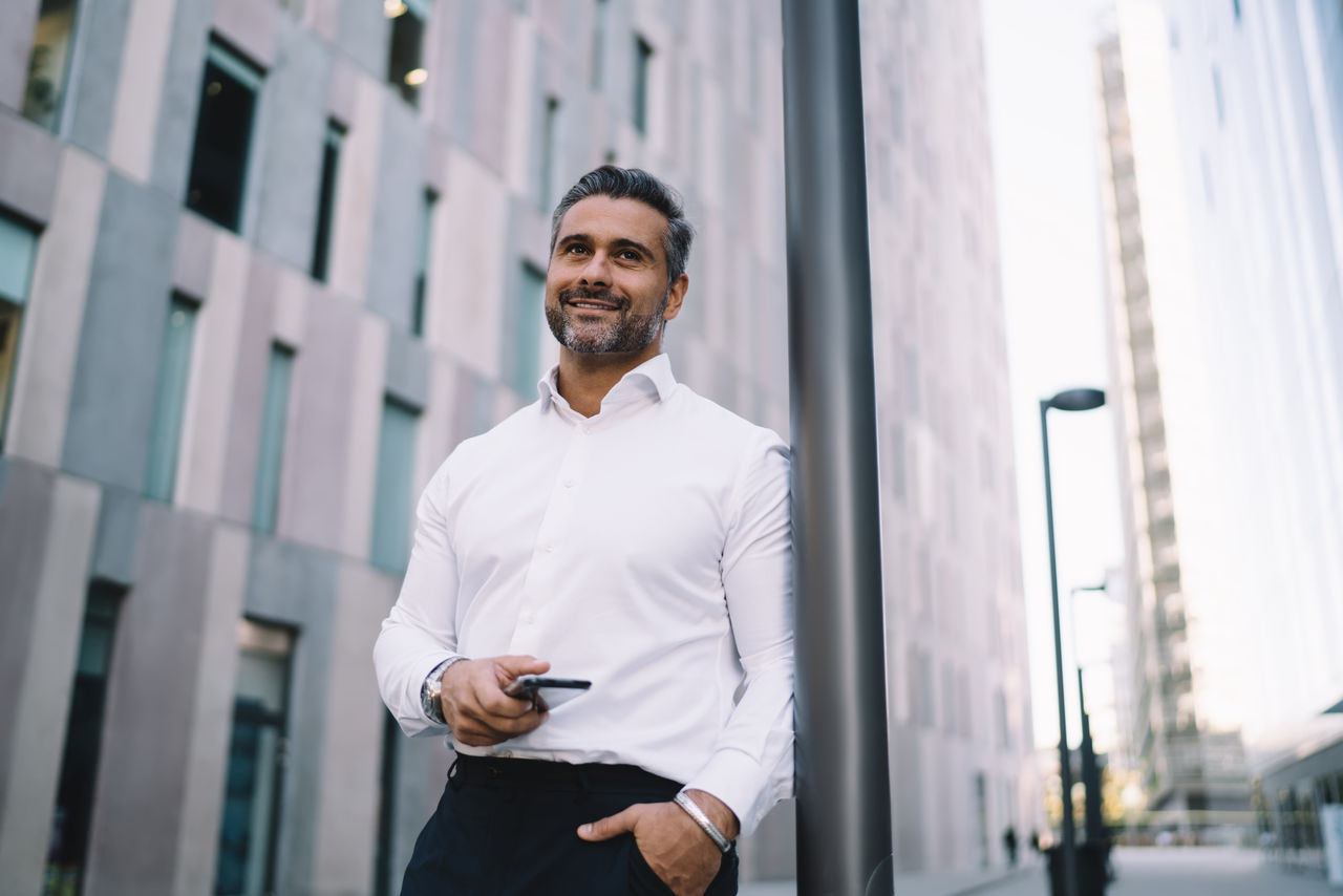Happy corporate director in formal white shirt holding cellphone gadget in hand waiting in financial district, successful middle aged employee with smartphone technology smiling in downtown