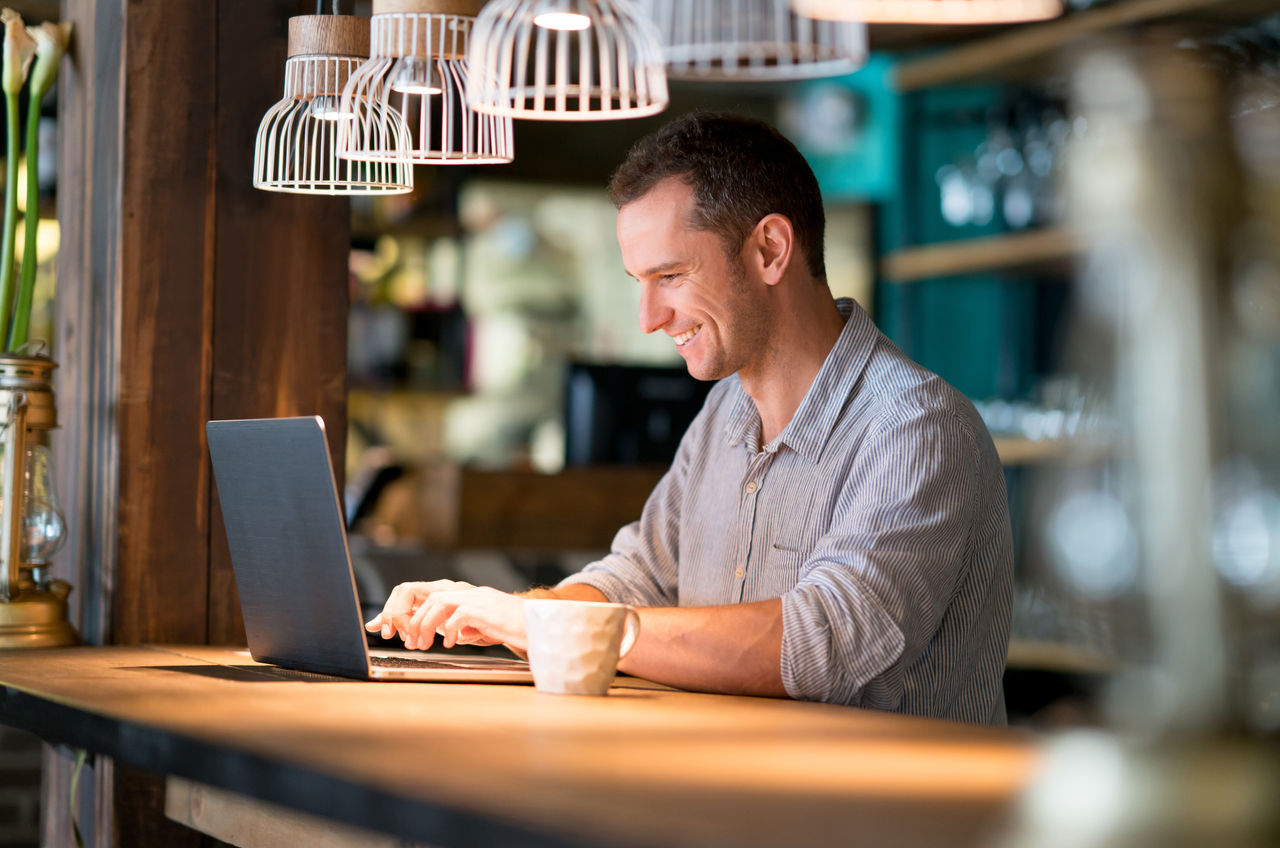 Portrait of a happy man working online at a cafe and drinking a cup of coffee